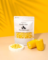 A package of Pookie Pets Freeze-Dried Corn Kernels with a transparent window showing the dried corn inside. The package is displayed alongside fresh corn cobs and a dish with scattered corn kernels.